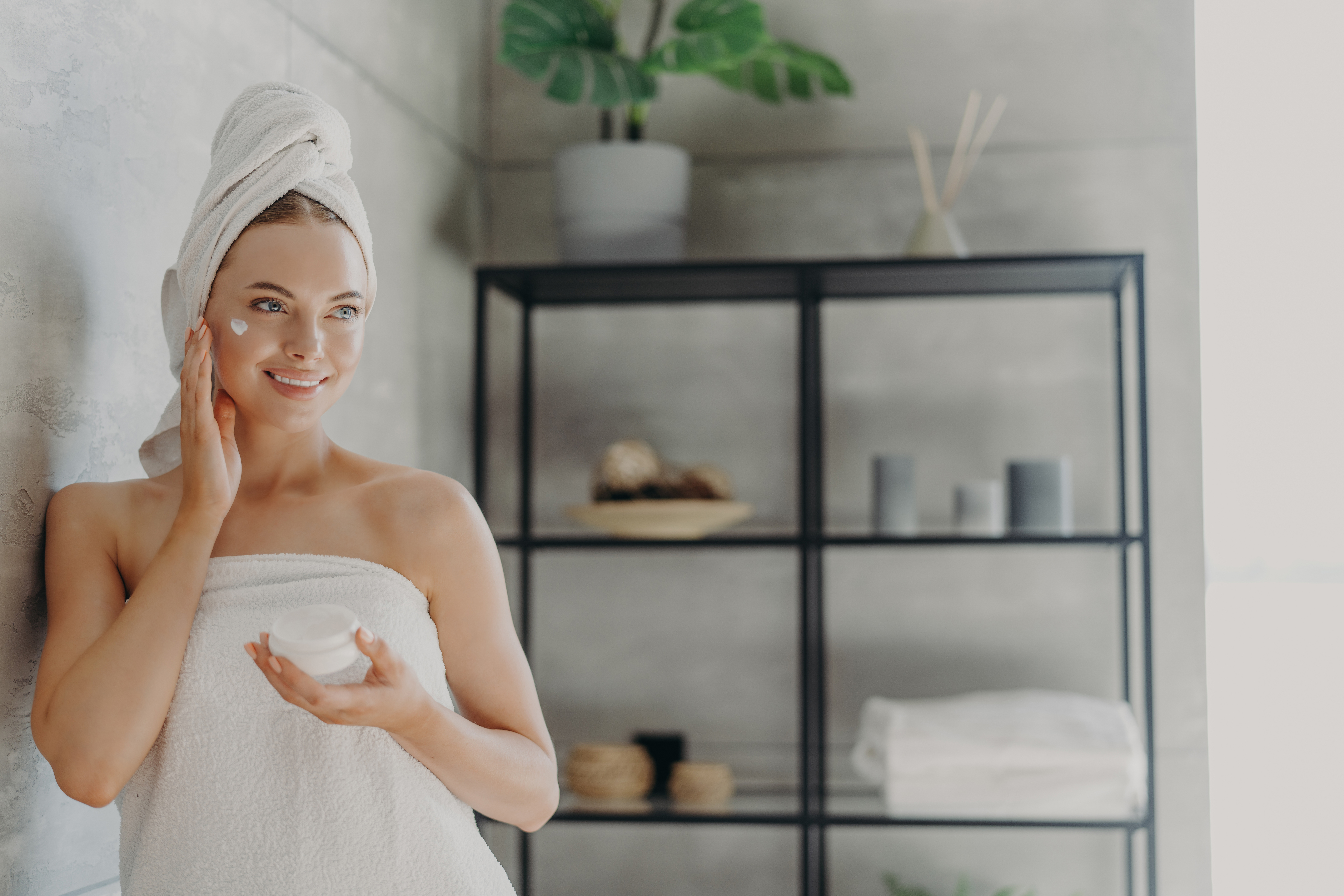 Lovely smiling Caucasian woman applies beauty cream on cheek, enjoys morning domestic skin care